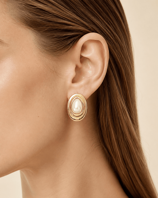 Close-up of a model wearing layered oval gold-tone stud earrings with a pearl-effect teardrop center
