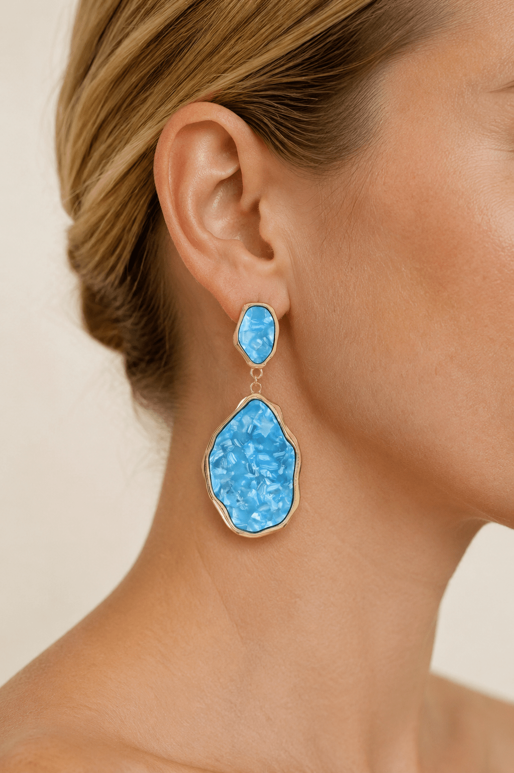 Close-up of a model wearing abstract gold-tone statement drop earrings with aqua stone-effect panels