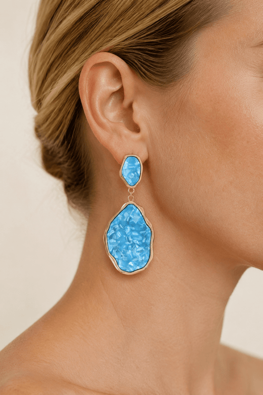 Close-up of a model wearing abstract gold-tone statement drop earrings with aqua stone-effect panels